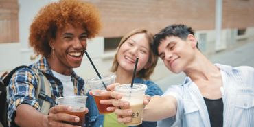 Three friends cheer with iced drinks, smiling joyfully.
