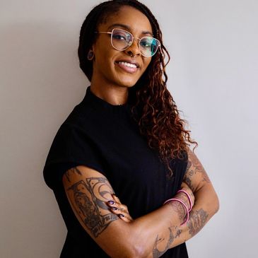 Smiling woman with glasses and tattoos, arms crossed confidently.