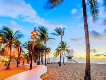 Sunset at a palm-lined beach promenade with a lifeguard tower and city buildings.