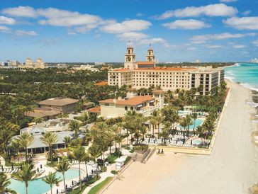 Aerial view of a luxurious beachfront resort with pools, palm trees, and clear blue ocean under a partly cloudy sky.
