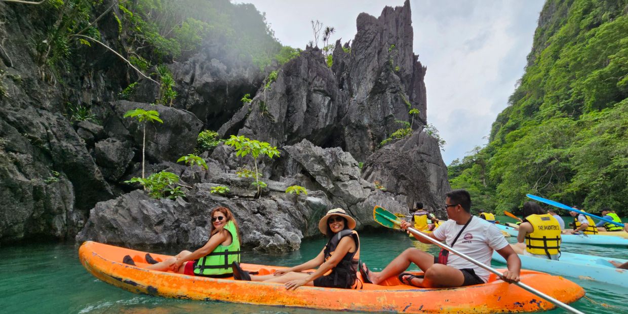 Three people kayaking near rocky cliffs and lush greenery.