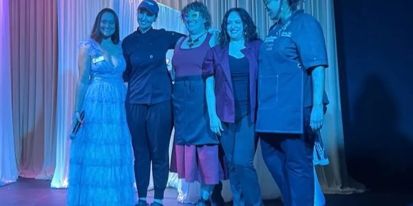 Five women standing together on stage under blue lighting.