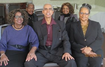 Five smiling elderly people posing together indoors in formal attire.