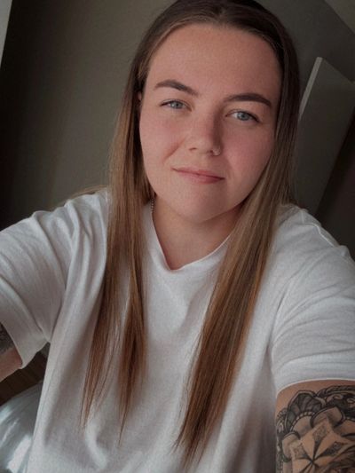 A young woman with long hair and tattoos wearing a white T-shirt.