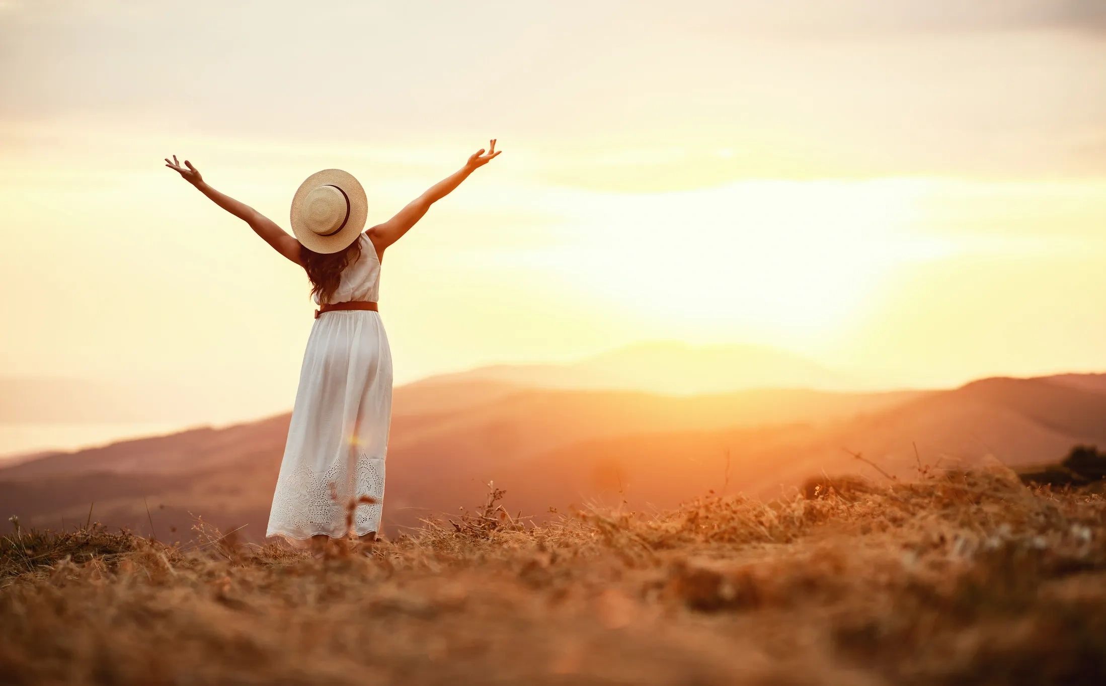Woman in white dress with hat enjoying a sunset on a hill.