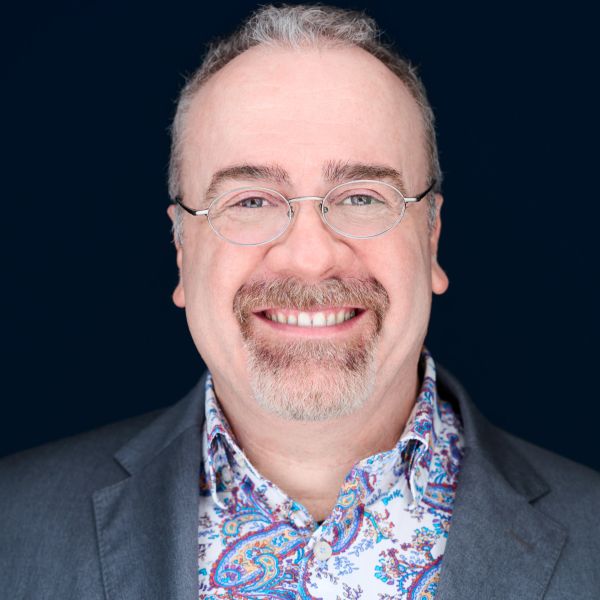 Smiling man with glasses wearing a paisley shirt and gray blazer.