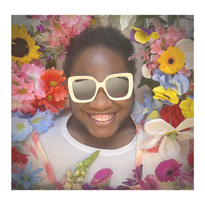 Brittany is laying down, head surrounded by bright colored flowers, wearing sunglasses and smiling