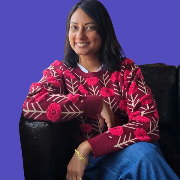 A woman smiles warmly, wearing a red floral sweater and blue jeans, seated on a black chair.