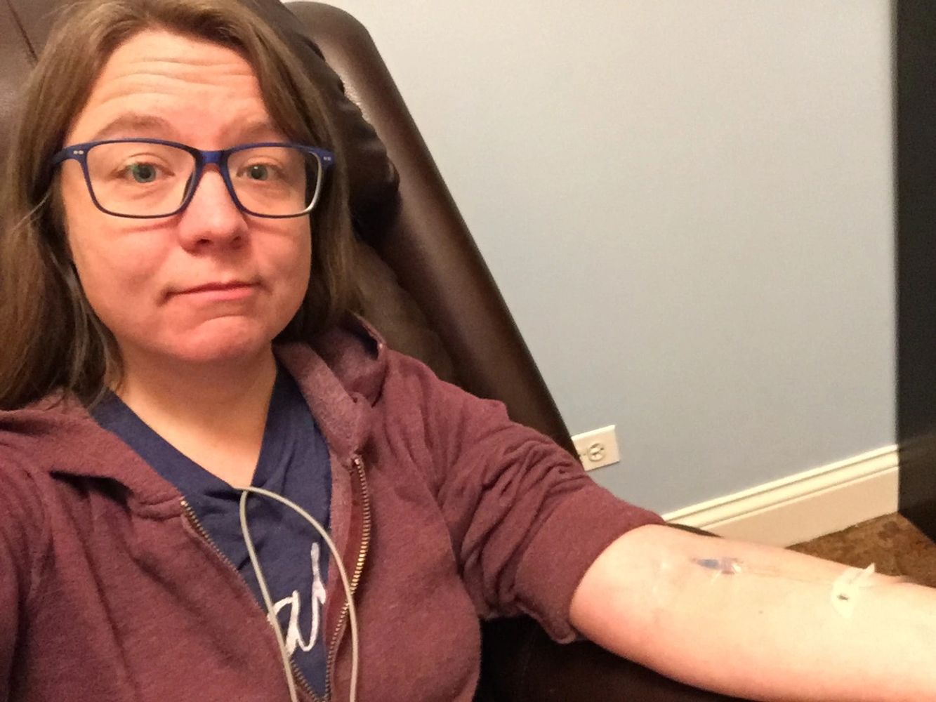 A white woman in a hoodie sits with an IV in her left arm. Wires come from her shirt.
