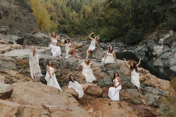 Group of women in white dresses posing on rocky terrain near water.