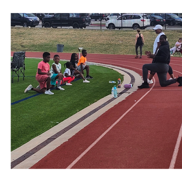 Coach kneels on track training young runners sitting on grass beside track.