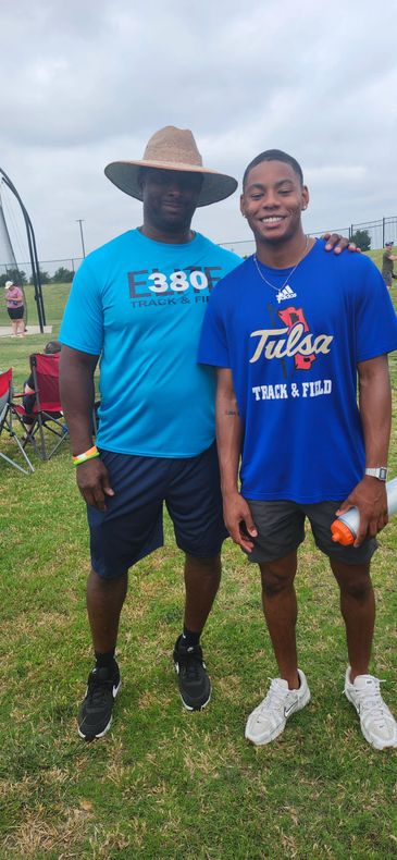 Two men posing outdoors in track and field attire, one wearing a wide-brimmed hat.
