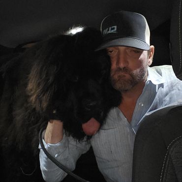 Man in a cap hugs a large black dog in a dimly lit setting.