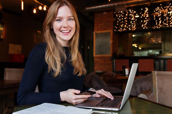 A blonde woman with her laptop computer and cell phone, sitting at a cafe with lights behind her.