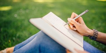 A green field, with a woman writing in a journal that is propped up on her knees.