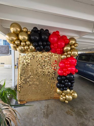 A decorative balloon arch in gold, black, and red with a gold sequin backdrop.