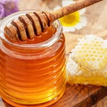 Jar of golden honey with a wooden honey dipper and honeycomb on a wooden board.