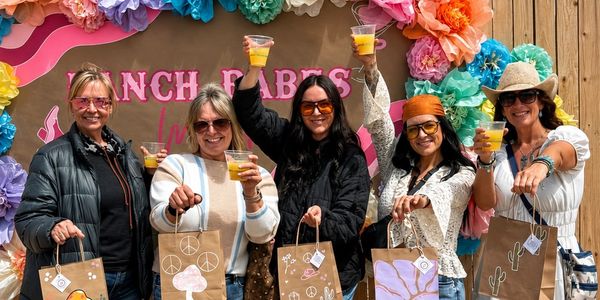 Group of five women holding drinks and gift bags at a colorful party.