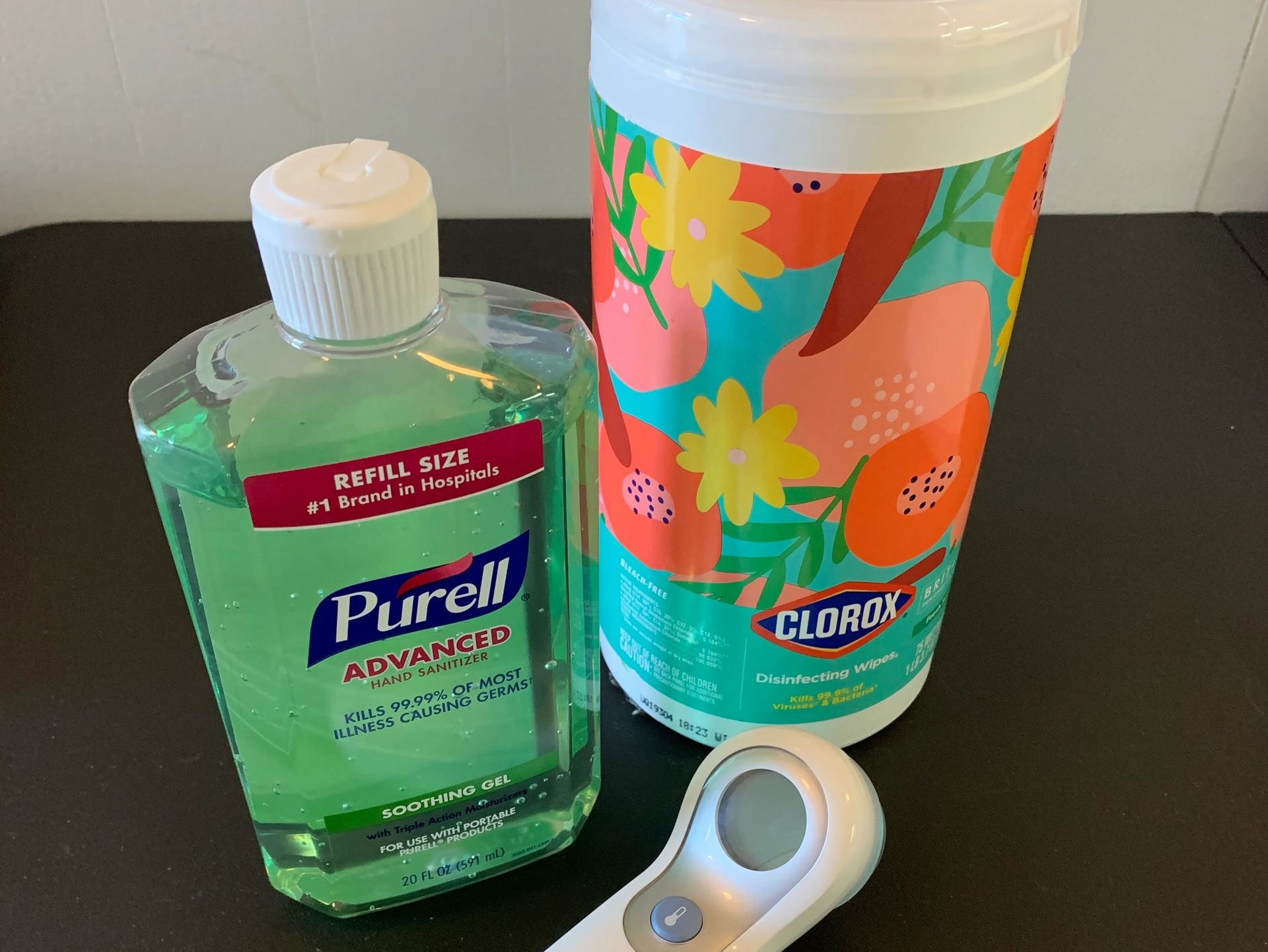 Purell hand sanitizer, Clorox disinfecting wipes, and a thermometer on a black surface.