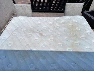 White mattress with visible stains placed outdoors near chairs and a gate.