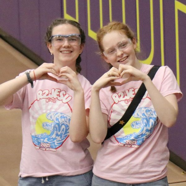 Two girls in matching pink shirts making heart signs with their hands.