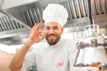 Smiling chef in white uniform making an OK gesture in a professional kitchen.