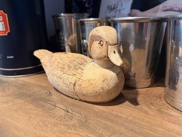 Intricately carved wooden duck figurine on a wooden surface with metal cups in the background.