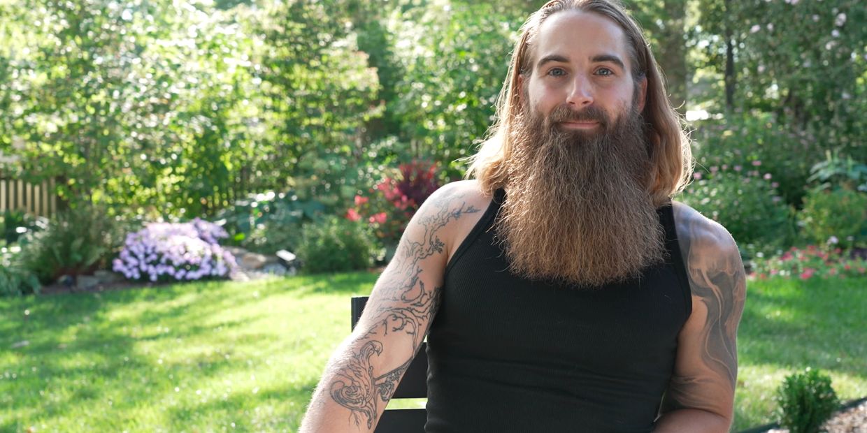 Bearded man with tattoos sits outdoors in a sunny garden.