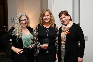 Three women smiling and holding wine glasses at a social event indoors.