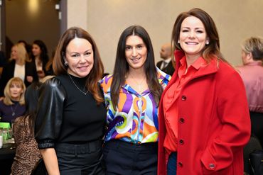 Three women smiling at a social event, dressed in vibrant and stylish outfits.