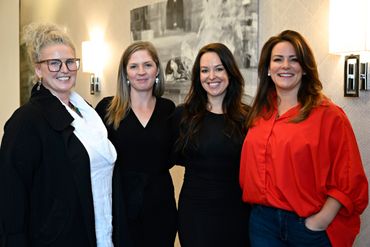 Four women standing together indoors, smiling warmly at the camera.