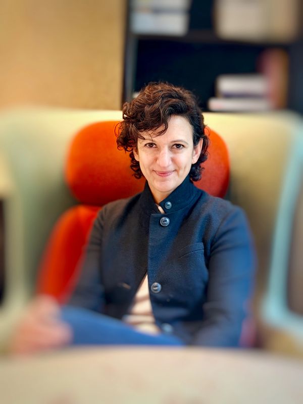 Woman with short curly hair sits confidently in an orange chair.