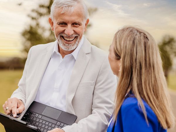 Smiling older man showing something on a laptop to a woman outdoors.
