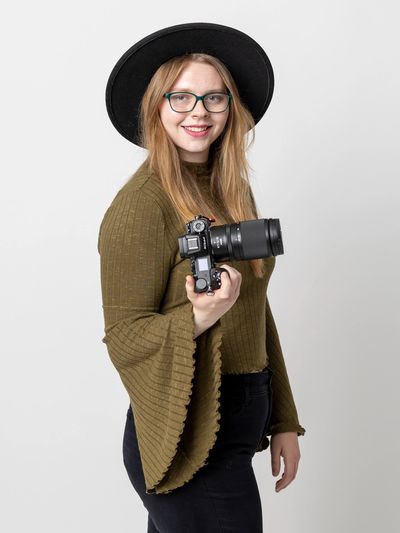 Young woman with glasses and a black hat holding a camera and smiling.