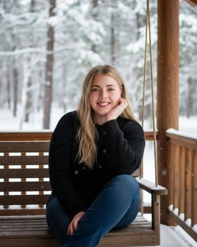 Smiling woman in black sweater sitting on a wooden swing amidst a snowy winter landscape.