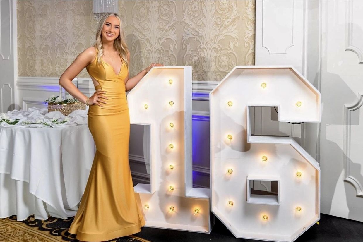 Young woman in yellow gown posing beside illuminated '16' sign.