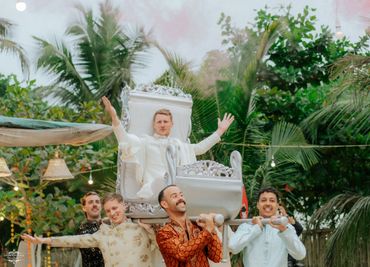 A man in white traditional attire is being carried on a decorated chair by four men in festive clothing.