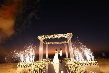 Couple standing under a floral arch at a beach wedding with fireworks at sunset.