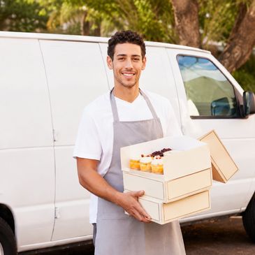 Smiling man in apron holding boxes of cupcakes outside a delivery van.