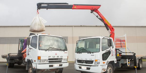 Two white trucks with a crane lifting a large bag outdoors.