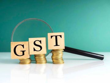 Wooden blocks spelling GST on stacks of coins with a magnifying glass.