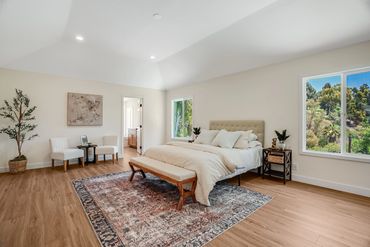 Spacious bedroom with a large bed, two chairs, and big windows overlooking greenery.