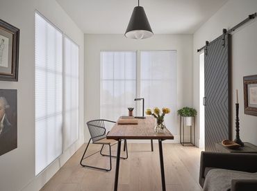 Minimalist home office with wooden desk, black chair, and natural light through covered windows.