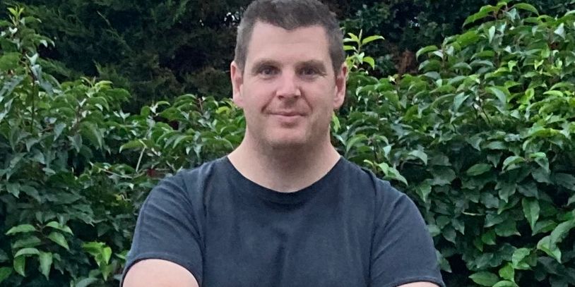Man in a black t-shirt standing outdoors with green leafy background.