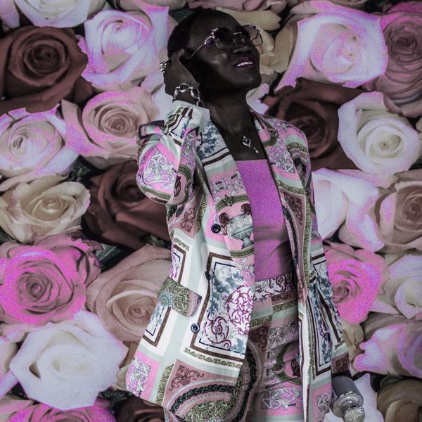 Person in patterned suit with microphone, standing against a rose wall.