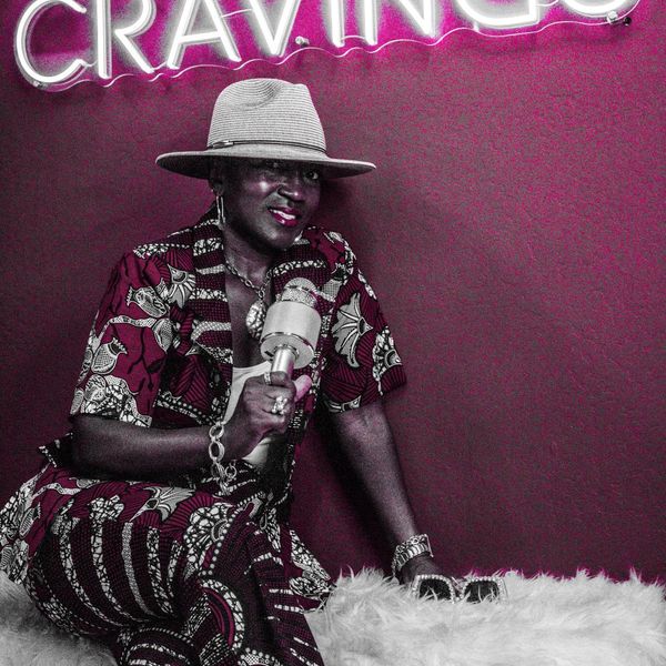 Woman in patterned outfit holding a microphone under a neon sign that says CRAVINGS.