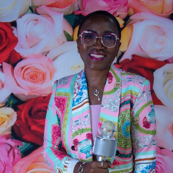 Woman in vibrant patterned suit holding a microphone, standing in front of a colorful rose backdrop.
