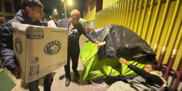 Persona entregando comida y otra con caja grande a un hombre en la calle por la noche.
