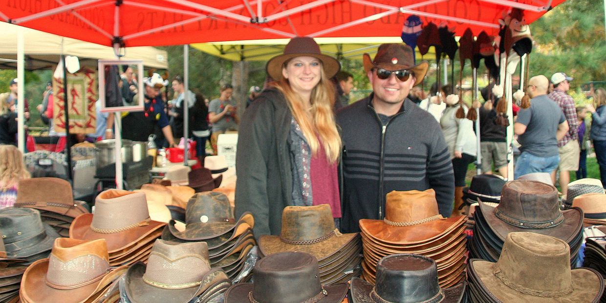 A man and woman smiling behind a table of stacked cowboy hats at an outdoor market.