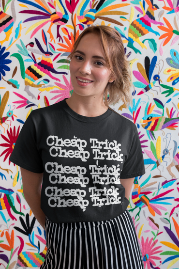 Smiling woman in a black 'Cheap Trick' t-shirt with colorful embroidered bird wall art behind her.